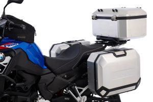 Wunderlich presentó el X-Case X55, el baúl de mayor tamaño de su línea X-Case. El mismo se caracteriza por una construcción de aluminio endurecido que combina resistencia y ligereza. Así se convierte en una opción ideal tanto para la aventura como para el turismo clásico, adaptándose perfectamente a las exigencias de los motociclistas que recorren largas distancias o que simplemente buscan un espacio seguro y confiable para transportar su equipaje. Diseño octogonal y aerodinámico La forma octogonal del X55 no es solo un detalle estético: permite una mayor durabilidad de la maleta y ayuda a reducir el peso total sin comprometer la robustez. Además, mejora la aerodinámica del conjunto, especialmente a altas velocidades, lo que contribuye a una mejor eficiencia y menor resistencia al viento durante la marcha. El top case viene equipado con una cerradura de cilindro de alta calidad y dos llaves. Por otro lado, el volumen de almacenamiento es de 55 litros, lo que alcanza para dos cascos, según el tamaño. El baúl tiene un volumen de 55 litros y una capacidad de carga de 10 kg. Fijación práctica y protección total Además, cuenta con un sistema de liberación rápida de acero inoxidable con asa de transporte integrada y plegable para abrir, cerrar y fijar al portaequipajes superior. Es 100 % resistente al agua y al polvo, lo que ofrece una máxima protección para el equipaje gracias a un sello de goma especial en todo su perímetro. También incorpora presillas de amarre en la tapa para asegurar equipaje adicional. El cilindro de cerradura está protegido contra la suciedad y la humedad mediante una tapa protectora. Por otro lado, el interior también fue rediseñado y ahora incorpora una espuma antideslizante que protege el contenido frente a las vibraciones y posibles golpes. Cierre personalizado Wunderlich permite a los usuarios personalizar el sistema de cierre. Los clientes pueden optar entre cerraduras codificadas, que permiten usar la misma llave del vehículo, o cilindros suministrados por dos llaves. Ambas opciones cuentan con una tapa protectora para preservar el mecanismo de la humedad y la suciedad.